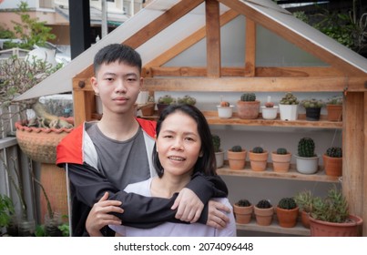 Mother And Son, Asian Teen, Black Hair. Mother And Child Concept, Hugging Each Other, Smiling Happily Looking At The Camera. At The Front Garden