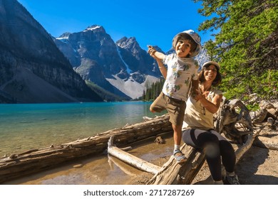 Mother and son are having fun while posing on a log by the turquoise waters of moraine lake, surrounded by the majestic mountains of banff national park on a sunny day