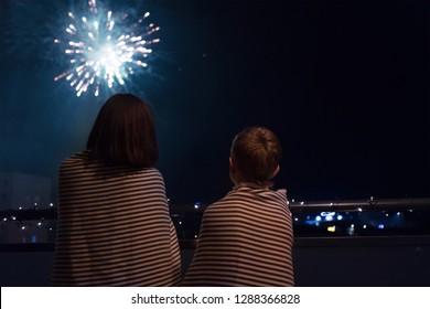 Mother and son looking at New Year celebration fireworks in night sky warmly wrapped in striped plaids standing on home balcony.