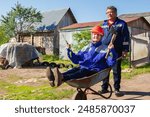 Mother and son. Man is driving an elderly woman in a construction helmet with an axe and a saw on a wheelbarrow. Concept of family gardening.