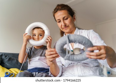 Mother And Son On The Sofa At Home Looking On Homemade Craft Decoration - Caucasian Boy Playing Silly While Mature Woman Work At Her Room - Real People Bonding Learning And Art Concept Front View