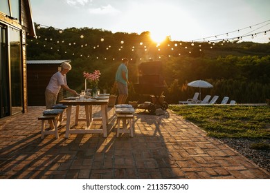 Mother And Son Preparing Dinner In The Backyard. Senior Woman Arranging Dining Table While Young Adult Man Barbecuing Beside