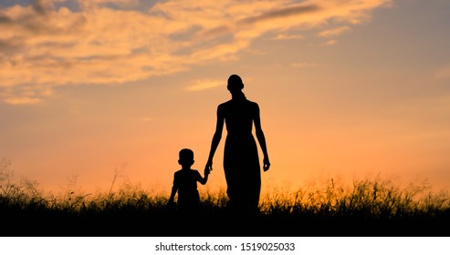 Mother And Son Silhouette Holding Hands Walking In A Grass Field At Sunset. 