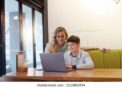 Mother And Son Sitting In A Coffee Shop, Doing Online Studying Or Homework Together On A Laptop, Mom Helping Her Son