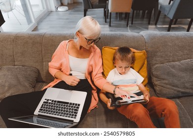 Mother and son sitting together on the couch. Woman, mother doing work on her laptop, working from home, while young boy sitting next to her doing schoolwork, homework on digital tablet. - Powered by Shutterstock