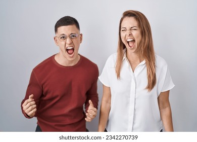 Mother and son standing together over isolated background angry and mad screaming frustrated and furious, shouting with anger. rage and aggressive concept. 