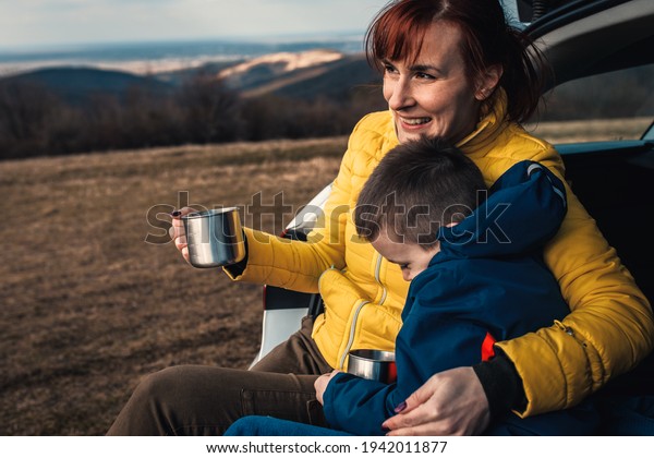 Mother and son take break from driving\
sitting by the car while drinking tea in\
nature.