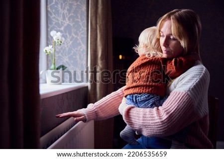 Mother With Son Trying To Keep Warm By Radiator At Home During Cost Of Living Energy Crisis Foto stock © 
