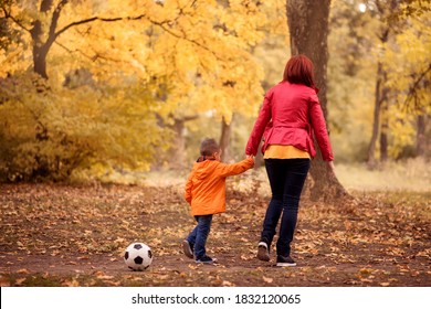 Mother And Son Walk Holding Hands In Autumn Park. Soccer Ball Laying Forgotten Behind Them. Mom Taking Toddler Kid Back Home After Football Training Or Game Outdoors. View From Behind. Copy Space