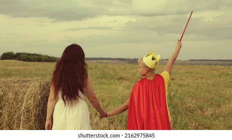 Mother Son Walk Through Green Field Sunset. Child Teenager Plays Superhero Nature. Hold Woman Hand. Teamwork. Happy Family. Fancy Dress Red Cloak, Crown, Sword. Woman Mother With Boy Son Travels Park