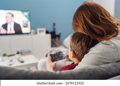 Mother And Son Young Woman And Her Boy Child Caucasian Kid Sitting On Sofa Bed At Home Making Video Call Or Playing Video Games On Mobile Smart Phone And Watch Tv During Quarantine Lockdown Back View