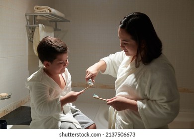 Mother Squeezing Tooth Paste On Toothbrush On Little Son When They Are Standing In Bathroom