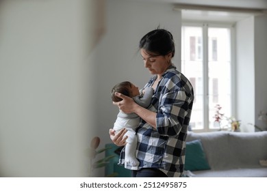 Mother standing in a bright living room cradling her newborn baby, who is wearing a cozy outfit and resting in her arms, with natural light pouring in through the window behind them. - Powered by Shutterstock