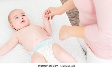 Mother Standing Next To Bed With Her Newborn Baby Boy Lying On His Back. Baby Boy On A Bed Wearing A Nappy Looking At The Mother And Smiling.