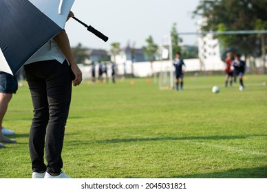 Mother Standing And Watching Her Son Playing Football In A School Tournament On A Clear Sky And Sunny Day. Sport, Active Lifestyle, Happy Family And Soccer Mom Concept.