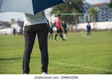 Mother Standing And Watching Her Son Playing Football In A School Tournament On A Clear Sky And Sunny Day. Sport, Active Lifestyle, Happy Family And Soccer Mom Concept.