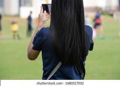 Mother Standing, Watching And Taking Pictures Of Her Daughter Playing Football In A School Tournament On A Clear Sky And Sunny Day. Sport, Active Lifestyle, Happy Family And Soccer Mom Concept.