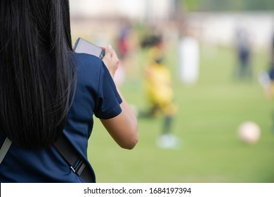 Mother Standing, Watching And Taking Pictures Of Her Daughter Playing Football In A School Tournament On A Clear Sky And Sunny Day. Sport, Active Lifestyle, Happy Family And Soccer Mom Concept.