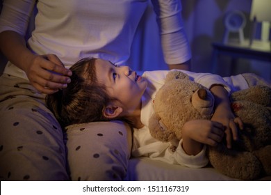 Mother Stroking Head Of Cute Girl Then Sleeping On Mom Lap On Bed In A Dark Bedroom At Night, Child Asian Girl Hug Teddy Bear, Comfortable Children At Home Concept