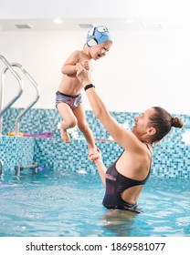 Mother Supporting Her Toddler Son In Swimming Pool Over Blue Water. Happy Kid Having Great Time With His Mom In Water, Wearing Swim Cap And Swimming Trunks. Concept Of Healthy Family