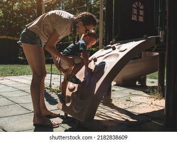 Mother Supporting And Holding Her 2 Years Old Child Son On Climbing Wall At Children Playground. Mom And Son Having Fun. Support Childhood Parenthood Symbol. Sunny, Summer Day.