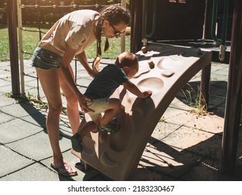 Mother Supporting And Holding Her 2 Years Old Child Son On Climbing Wall At Children Playground. Mom And Son Having Fun. Support Childhood Parenthood Symbol. Sunny, Summer Day.