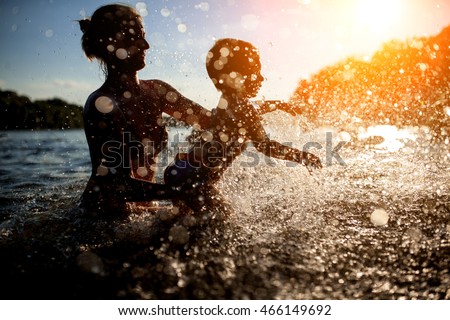 mother swim with baby in blue water at sunset; female with child bathe in lake or river and making water drops;