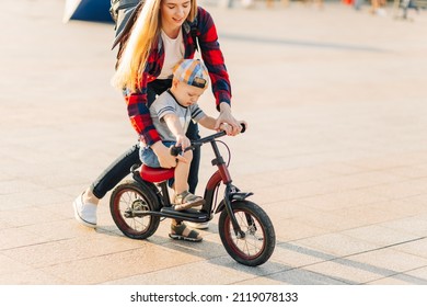 Mother Takes Care Of A Child, Training To Ride A Bicycle, Running After Her Son In A Public Park. A Child Smiling At Mom With A Happy Face Loves To Play Together. The Boy Feels Fun On The Bike.