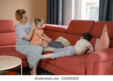 Mother Taking Care Of Two Kids At Home On Red Sofa. Making Hair For Daughter And Talking To Son In The Morning. Casual Family Moment. Interaction And Daily Routine With Children