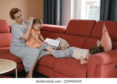 Mother Taking Care Of Two Kids At Home On Red Sofa. Making Hair For Daughter And Talking To Son In The Morning. Casual Family Moment. Interaction And Daily Routine With Children