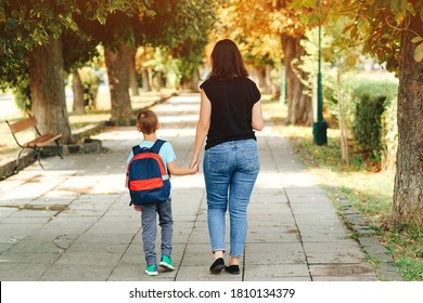 Mother Taking Child To School. First Day At School. Mother Leads A Little School Boy In First Grade. Parent And Pupil Of Primary School Go Hand In Hand. Woman And Kid With Backpack Behind The Back.