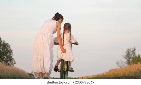 Mother teaches child girl daughter ride two-wheeled bicycle. happy family. Happy cute little girl learns ride with mom bike park sunset. sunshine park happy family. mom teaches daughter kid ride bike - Powered by Shutterstock