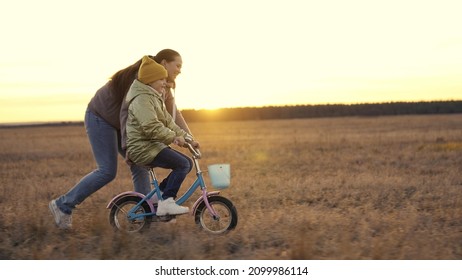 mother teaches little daughter ride two-wheeled bike at sunset, child pedals on bicycle in sunshine, outdoor outdoor activities, exercise at dawn travel, happy family life, childhood dream drive - Powered by Shutterstock