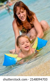 Mother Teaching Baby To Swim In Outdoor Pool. Child Is Learning To Swim With Inflatable Arm Floaties Sleeves On Vacation In The Summer On A Sunny Day. Swimming Teacher Or Instructor.