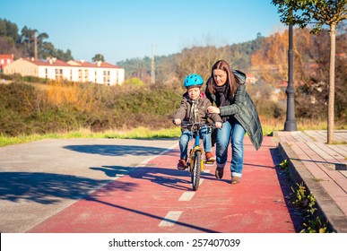 Mother Teaching Her Son To Ride A Bicycles In The City On A Sunny Winter Day.  Family Leisure Outdoors Concept.