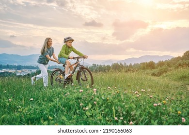 Mother Teaching Son To Ride Bicycle. Happy Cute Boy In Helmet Learn To Riding A Bike In Park On Green Meadow In Summer Day At Sunset Time. Family Weekend. 4K Video Slow Motion