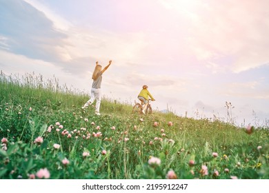 Mother Teaching Son To Ride Bicycle. Happy Cute Boy In Helmet Learn To Riding A Bike In Park On Green Meadow In Summer Day At Sunset Time. Family Weekend. 4K Video Slow Motion