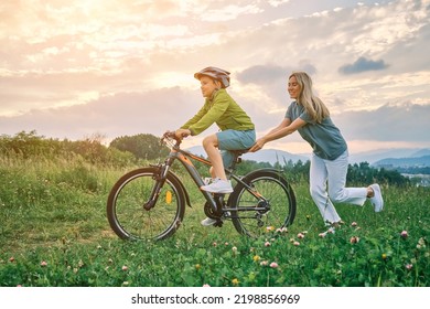 Mother Teaching Son To Ride Bicycle. Happy Cute Boy In Helmet Learn To Riding A Bike In Park On Green Meadow In Summer Day At Sunset Time. Family Weekend. 4K Video Slow Motion