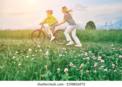 Mother Teaching Son To Ride Bicycle. Happy Cute Boy In Helmet Learn To Riding A Bike In Park On Green Meadow In Summer Day At Sunset Time. Family Weekend. 4K Video Slow Motion