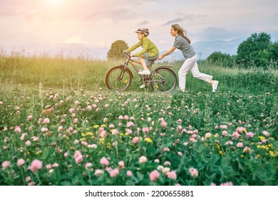 Mother Teaching Son To Ride Bicycle. Happy Cute Boy In Helmet Learn To Riding A Bike In Park On Green Meadow In Summer Day At Sunset Time. Family Weekend. 4K Video Slow Motion