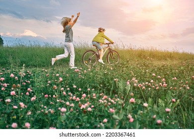 Mother Teaching Son To Ride Bicycle. Happy Cute Boy In Helmet Learn To Riding A Bike In Park On Green Meadow In Summer Day At Sunset Time. Family Weekend. 4K Video Slow Motion