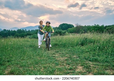 Mother Teaching Son To Ride Bicycle. Happy Cute Boy In Helmet Learn To Riding A Bike In Park On Green Meadow In Summer Day At Sunset Time. Family Weekend. 4K Video Slow Motion