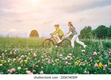 Mother Teaching Son To Ride Bicycle. Happy Cute Boy In Helmet Learn To Riding A Bike In Park On Green Meadow In Summer Day At Sunset Time. Family Weekend. 4K Video Slow Motion