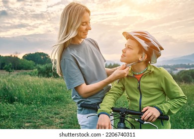 Mother Teaching Son To Ride Bicycle. Happy Cute Boy In Helmet Learn To Riding A Bike In Park On Green Meadow In Summer Day At Sunset Time. Family Weekend. 4K Video Slow Motion