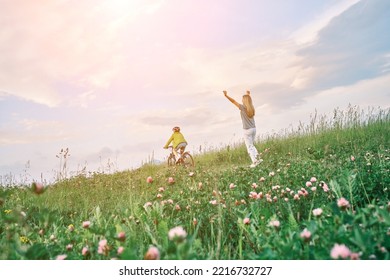 Mother Teaching Son To Ride Bicycle. Happy Cute Boy In Helmet Learn To Riding A Bike In Park On Green Meadow In Summer Day At Sunset Time. Family Weekend. 4K Video Slow Motion