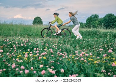 Mother Teaching Son To Ride Bicycle. Happy Cute Boy In Helmet Learn To Riding A Bike In Park On Green Meadow In Summer Day At Sunset Time. Family Weekend. 4K Video Slow Motion