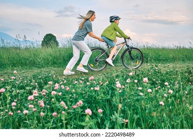 Mother Teaching Son To Ride Bicycle. Happy Cute Boy In Helmet Learn To Riding A Bike In Park On Green Meadow In Summer Day At Sunset Time. Family Weekend. 4K Video Slow Motion