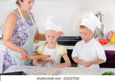 Mother Teaching Two Young Children, A Girl And Boy Dressed In White Chefs Uniforms With Aprons And Toques, To Bake In The Kitchen Showing Them How To Roll Out Dough For A Homemade Pizza Base