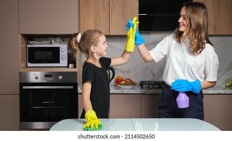 Mother And Teen Daughter With Colorful Gloves Clean White Table With Green Sponge And Cleanser Giving High Five In Kitchen At Home.