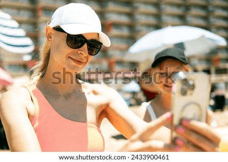 A mother and teenage daughter in swimsuits take a selfie against the backdrop of a summer beach on their phone, capturing cherished moments together.